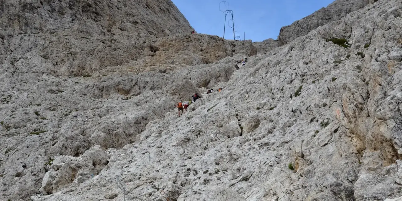 Escursionisti affrontano la **difficoltà** del sentiero verso il **Rifugio Re Alberto**, aggrappandosi a cavi d'acciaio su un ripido pendio roccioso.