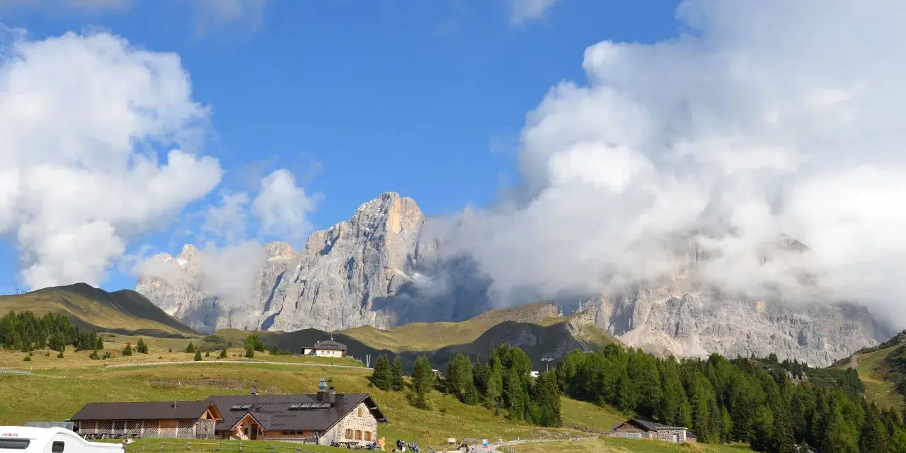 Panorami mozzafiato al Passo Rolle: malghe, boschi e vette imponenti sotto un cielo azzurro con nuvole bianche.