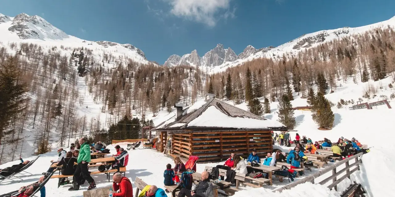 Malga Valcigolera, un rifugio alpino innevato con gente seduta ai tavoli. Per sapere come arrivare, guarda l'immagine.