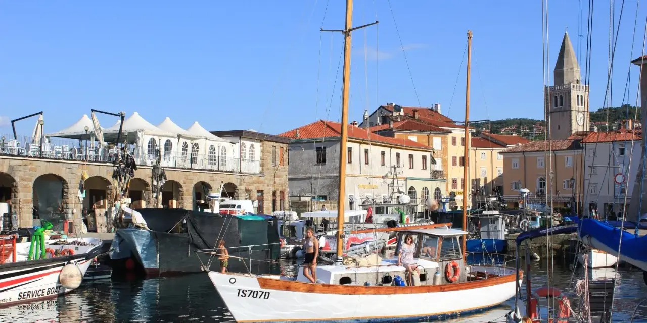 Porto di Muggia cosa vedere: barche a vela, edifici storici e un campanile svettano sotto il cielo azzurro.