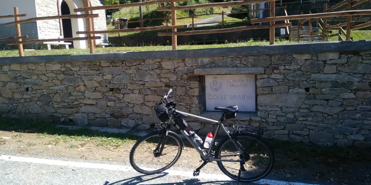 Bicicletta ferma sul ciglio della strada, con un muretto in pietra e un cartello che indica "Colle Braida".