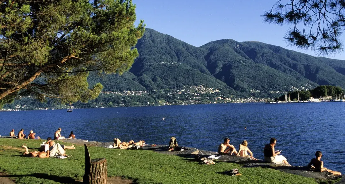 Persone rilassate sui prati e sulle rive di uno dei bellissimi laghi piemontesi, con montagne verdi sullo sfondo.