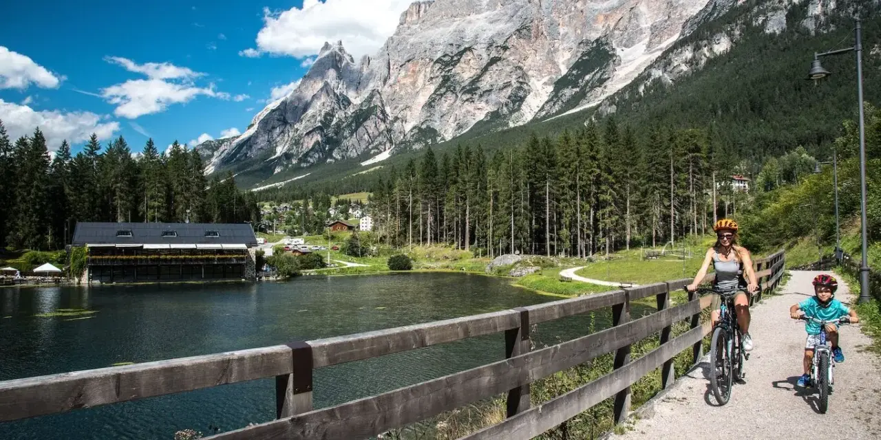 Famiglia in bicicletta sul ponte del **lago di San Vito**, con montagne imponenti e boschi sullo sfondo.