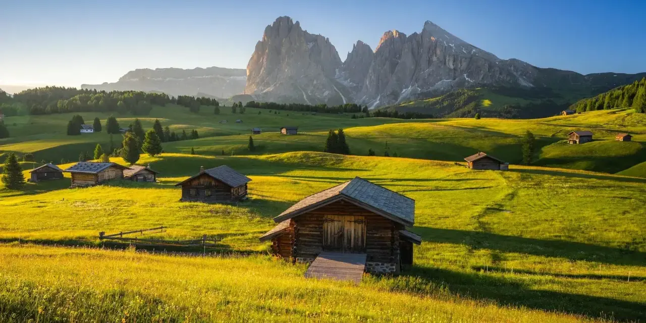 Alpe di Siusi al tramonto, con baite in legno su prati verdi e montagne imponenti sullo sfondo.