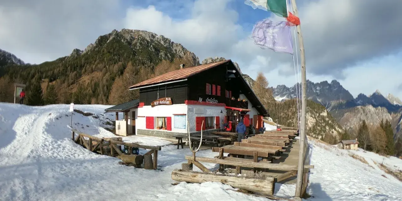 Rifugio Antelao innevato, con tavoli esterni e bandiere. Informazioni su come arrivare al rifugio sono visibili.