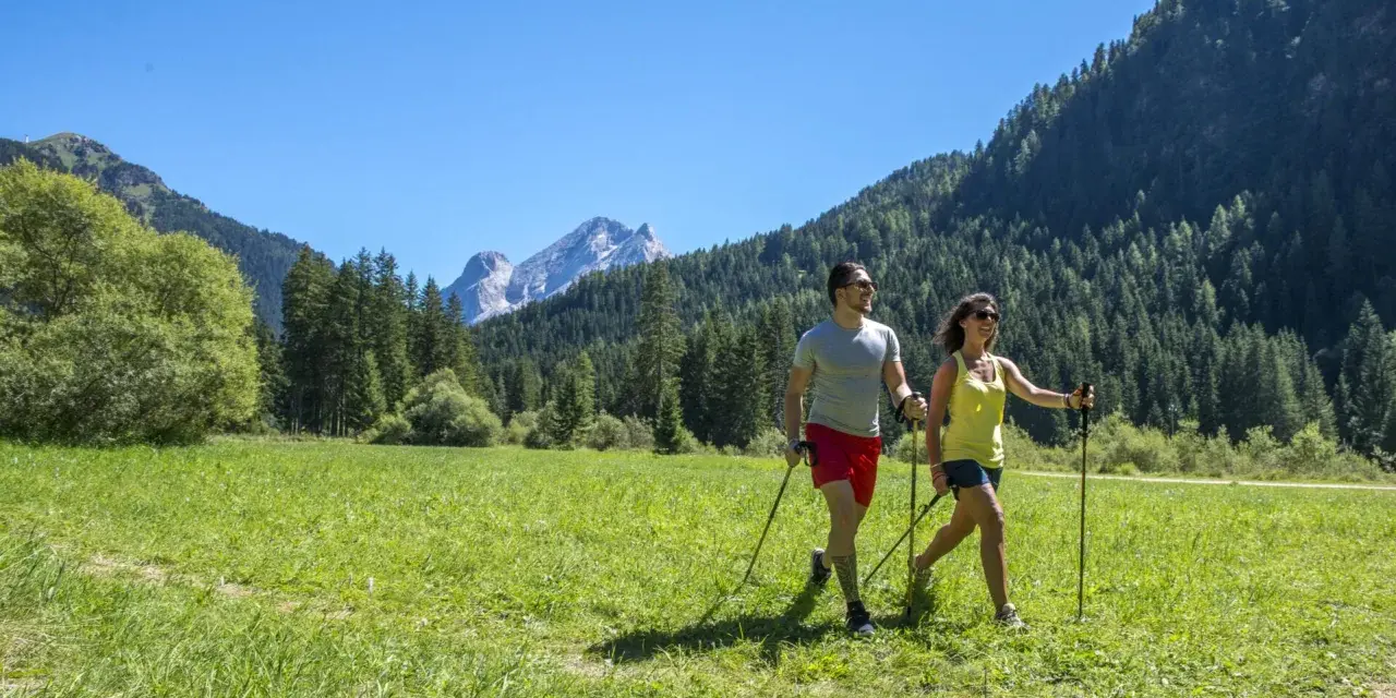 Coppia fa nordic walking in un prato verde con montagne sullo sfondo, ideale per canazei escursioni facili.