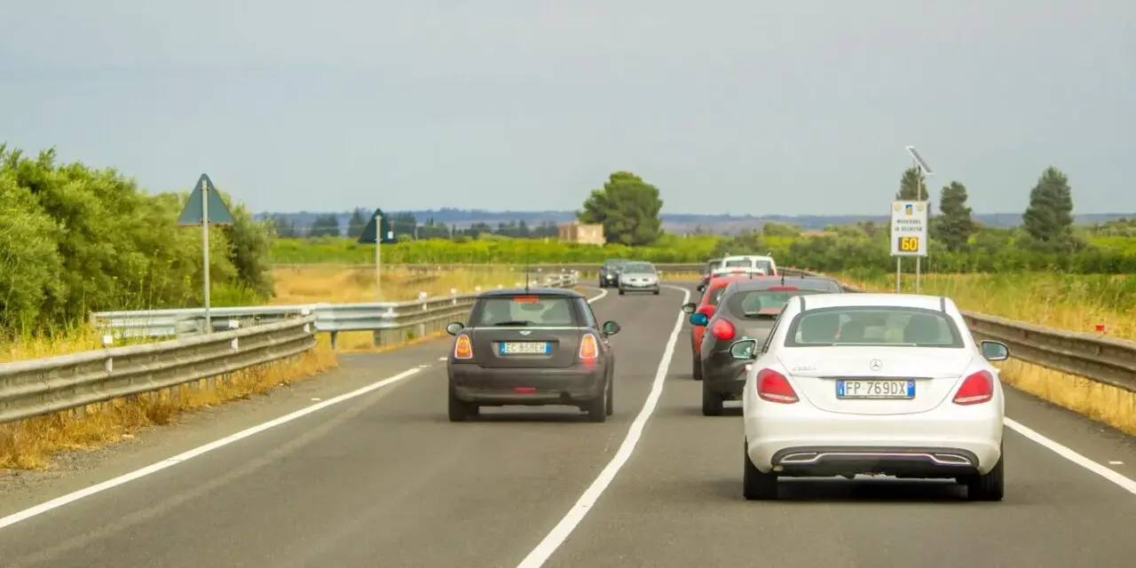 Traffico in colonna su strada con vegetazione ai lati. Un'auto tenta il sorpasso linea continua, mettendo a rischio la sicurezza.