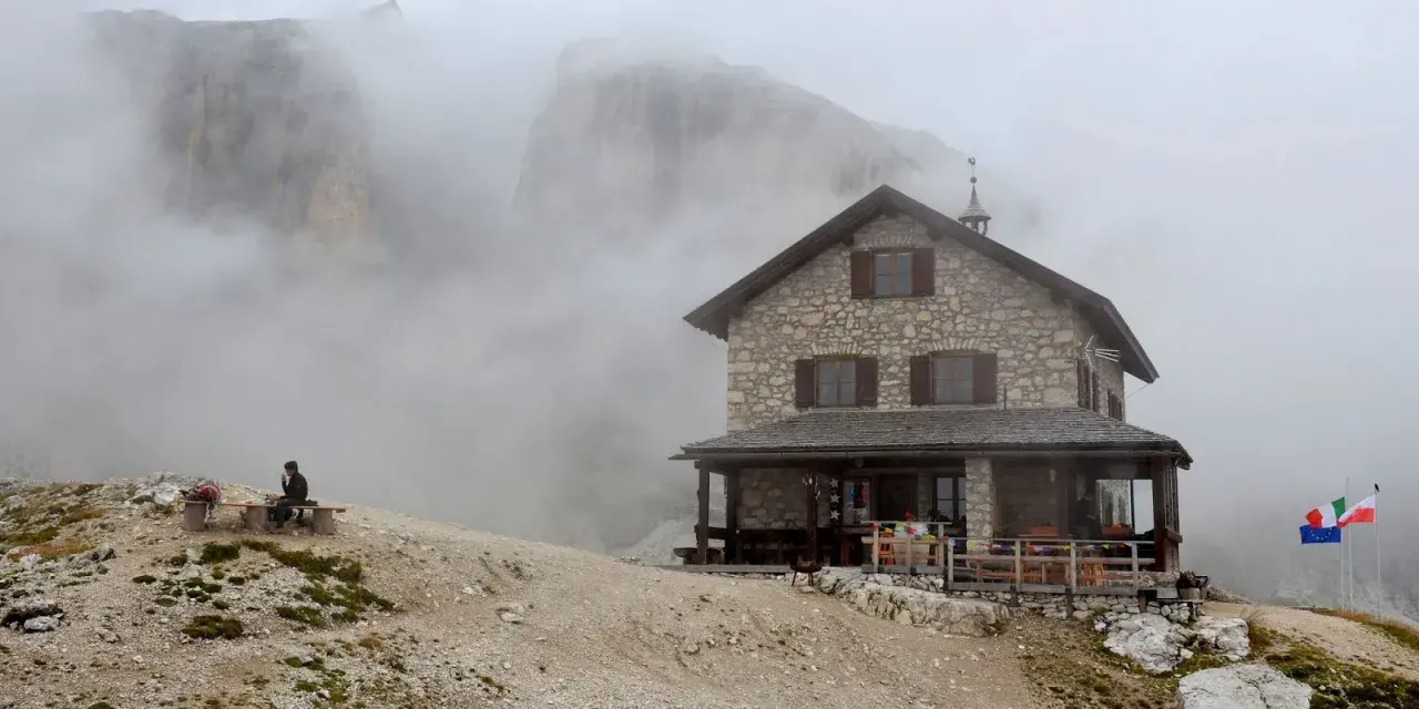 Rifugio Franz Kostner avvolto dalla nebbia, con un escursionista solitario su una panchina e le bandiere che sventolano.