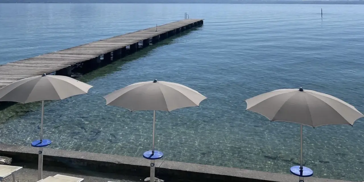 Ombrelloni e lettini al lido delle Bionde, con un pontile in legno che si estende sul lago limpido e montagne all'orizzonte.