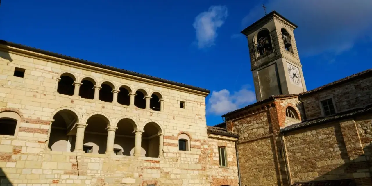Antica **cella monte** con portici ad archi e torre campanaria sotto un cielo azzurro.
