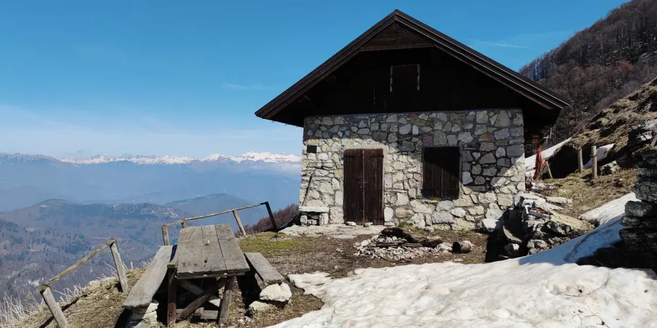 Bivacco Monte Grappa, rifugio in pietra con tavolo da picnic e vista sulle montagne innevate.