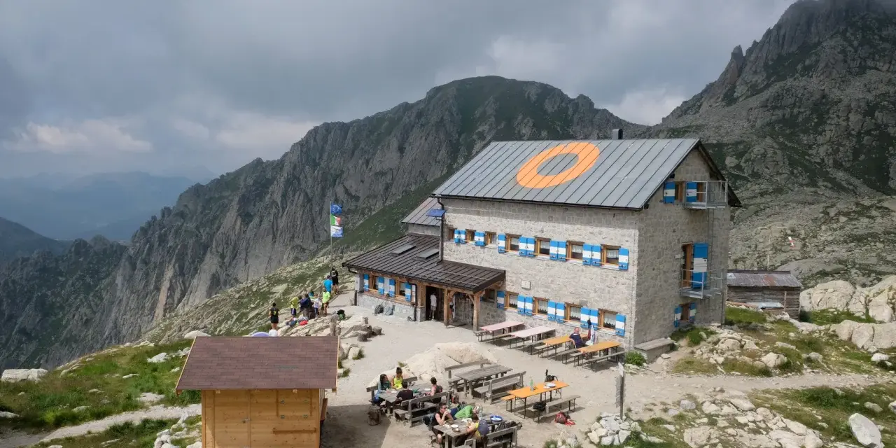 Il rifugio Cima d'Asta, con le sue finestre blu e arancioni, accoglie escursionisti tra rocce e montagne nebbiose.