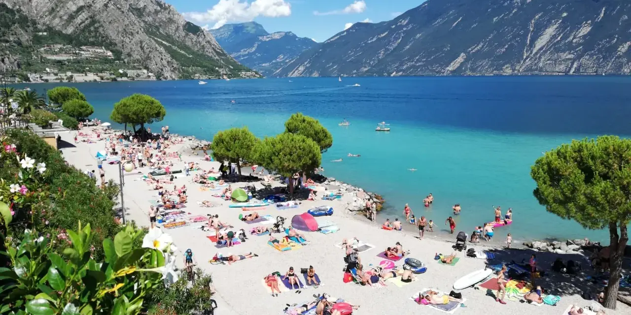 Una vivace spiaggia di Limone sul Garda, con gente che prende il sole e nuota nelle acque turchesi del lago.