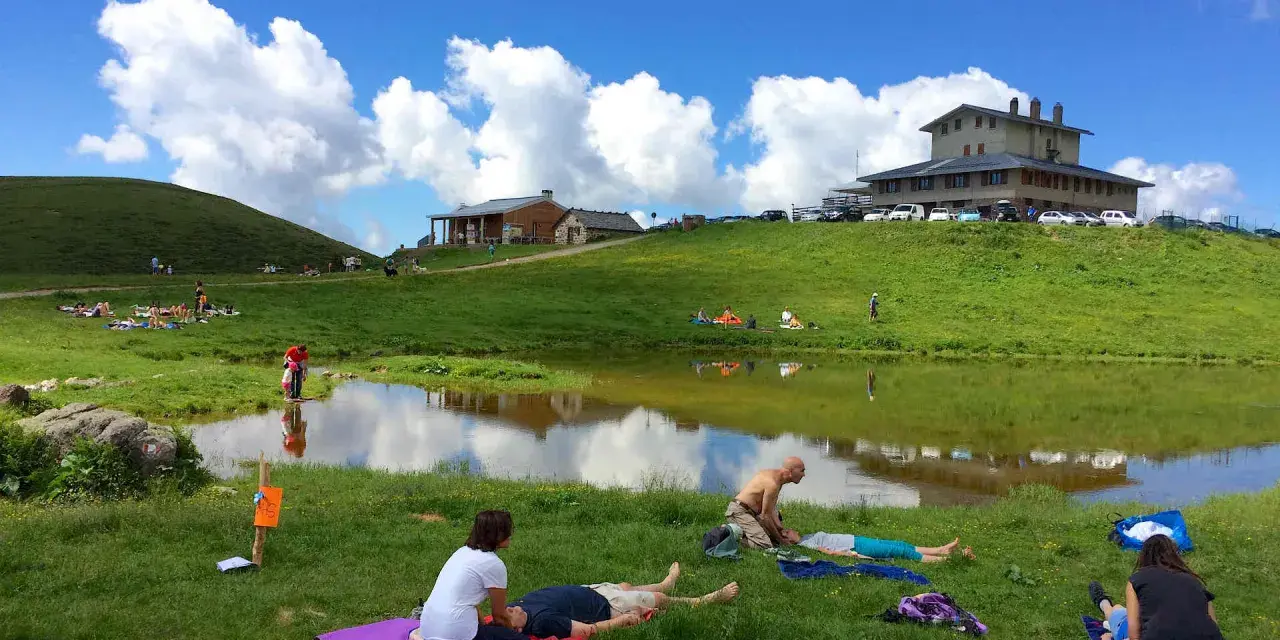 Persone rilassate sui prati verdi, vicino a un lago, sotto un cielo azzurro con nuvole bianche. Sembra un luogo ideale per i piani dell'avaro, godendosi la natura.
