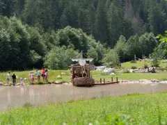 Bambini giocano vicino a un galeone di legno in un lago, mentre altri si rilassano sull'erba. Perfetto per le **san vigilio di marebbe passeggiate** estive.