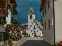 Un pittoresco vicolo a Rocca Pietore, con case fiorite e una chiesa dal cielo tempestoso.