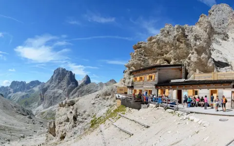 Rifugio alpino con gente seduta all'esterno, circondato da rocce imponenti e montagne. Un luogo ideale per le 12 escursioni da fare in Val di Fassa.