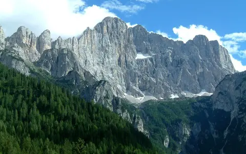 Imponente parete rocciosa del monte Civetta, con foreste di conifere ai suoi piedi e nuvole bianche nel cielo azzurro.