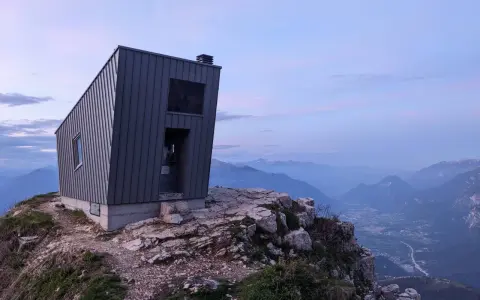 Il bivacco Giacomelli alla Madonnina, una struttura moderna su una cresta rocciosa, domina un paesaggio montano al tramonto.