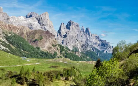 Strada panoramica verso le Pale di San Martino, con verdi pascoli e boschi che conducono alle maestose vette rocciose.