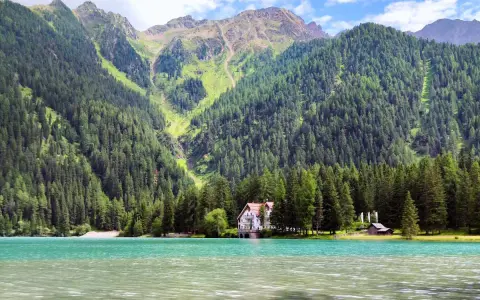 Un incantevole **Antholzer See** con acque turchesi, circondato da fitti boschi e imponenti montagne. Una casa bianca e un piccolo rifugio si affacciano sul lago.