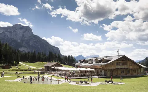 Un rifugio alpino con tavoli all'aperto e un laghetto dove giocano bambini, incorniciato da boschi e montagne imponenti sui **piani di Pezzè**.
