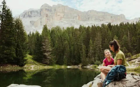 Mamma e figlia sedute su una roccia vicino a un lago alpino, con uno zaino blu. Un momento di relax per il loro lech dla lunch, con montagne imponenti sullo sfondo.