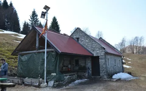 Bivacco Vallon Scuro: rifugio in pietra con tetto rosso, pannelli solari e telo verde. Un uomo si ripara dal freddo.