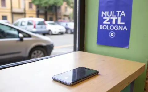 Un cellulare su un tavolo, con un cartello "Multa ZTL Bologna" appeso al muro. Fuori, auto in transito.