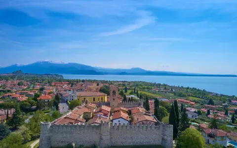 Vista aerea di Moniga del Garda, con il castello medievale in primo piano, il lago e le montagne sullo sfondo.