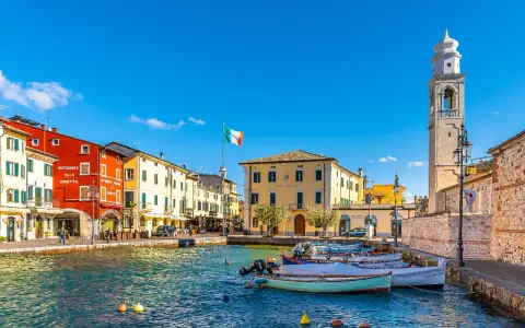Un pittoresco porto dove si trova Lazise, con case colorate, barche e un campanile svettante sotto un cielo azzurro.