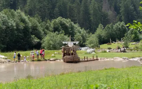 Bambini giocano vicino a un galeone di legno in un lago, mentre altri si rilassano sull'erba. Perfetto per le **san vigilio di marebbe passeggiate** estive.