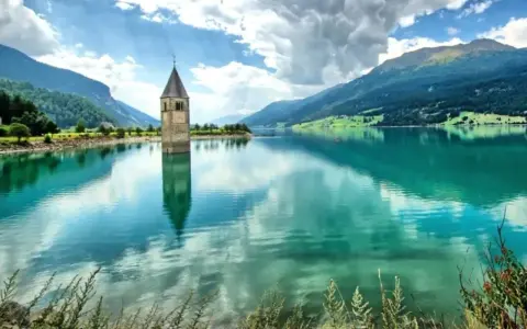 Torre sommersa nel lago di Resia, con montagne verdi e cielo nuvoloso.