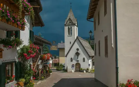 Un pittoresco vicolo a Rocca Pietore, con case fiorite e una chiesa dal cielo tempestoso.