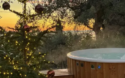 Un albero di Natale illuminato e una vasca idromassaggio fumante al tramonto sul Lago di Garda. Il camping è aperto tutto l'anno, un'oasi di relax invernale.