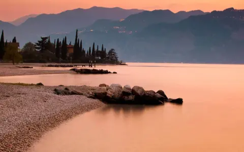 Brenzone sul Garda: cosa vedere? Spiaggia di ciottoli, cipressi e montagne al tramonto sul lago.
