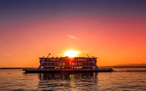 Traghetto in navigazione sul Lago di Garda al tramonto, con il sole che illumina il cielo e il riflesso sull'acqua.