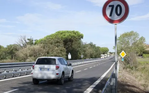 Un'auto bianca percorre una strada extraurbana con limite di 70 km/h. Alberi e cielo azzurro fanno da cornice.