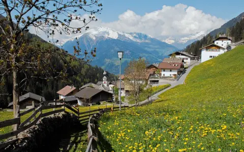 Un prato fiorito di gialli ranuncoli abbraccia San Leonardo in Passiria, con case tradizionali e montagne innevate sullo sfondo.