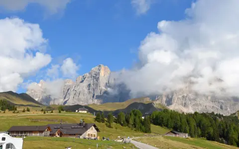 Panorami mozzafiato al Passo Rolle: malghe, boschi e vette imponenti sotto un cielo azzurro con nuvole bianche.