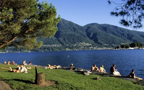 Persone rilassate sui prati e sulle rive di uno dei bellissimi laghi piemontesi, con montagne verdi sullo sfondo.