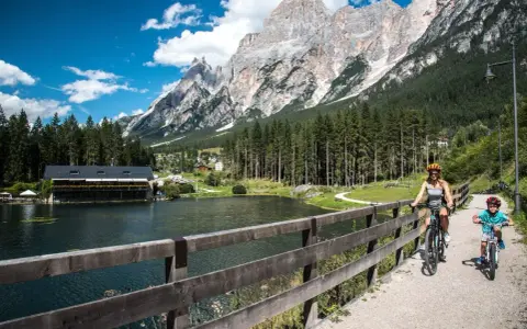 Famiglia in bicicletta sul ponte del **lago di San Vito**, con montagne imponenti e boschi sullo sfondo.