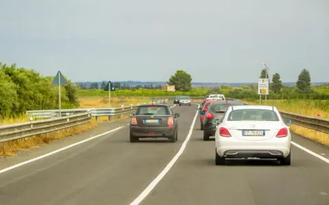 Traffico in colonna su strada con vegetazione ai lati. Un'auto tenta il sorpasso linea continua, mettendo a rischio la sicurezza.