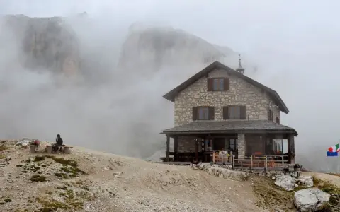 Rifugio Franz Kostner avvolto dalla nebbia, con un escursionista solitario su una panchina e le bandiere che sventolano.