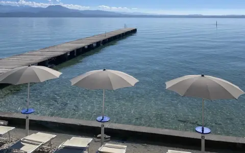 Ombrelloni e lettini al lido delle Bionde, con un pontile in legno che si estende sul lago limpido e montagne all'orizzonte.