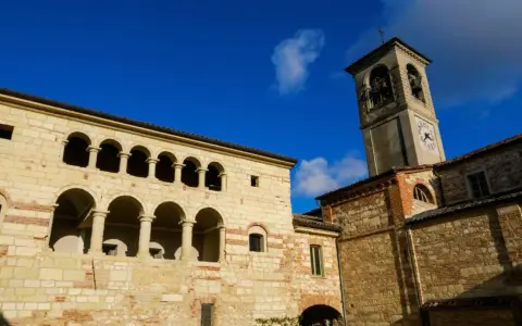 Antica **cella monte** con portici ad archi e torre campanaria sotto un cielo azzurro.