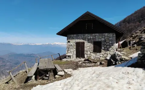 Bivacco Monte Grappa, rifugio in pietra con tavolo da picnic e vista sulle montagne innevate.