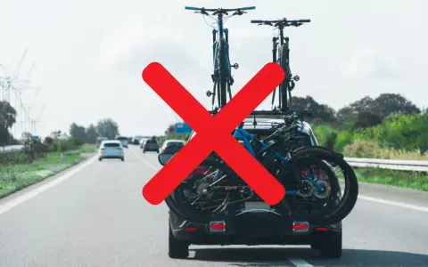 Attenzione al carico sporgente auto! Biciclette sul portabici posteriore, ma una grande X rossa indica un problema di sicurezza.