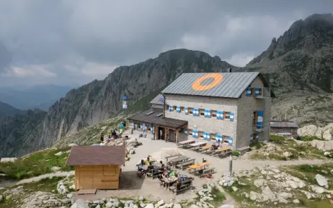 Il rifugio Cima d'Asta, con le sue finestre blu e arancioni, accoglie escursionisti tra rocce e montagne nebbiose.