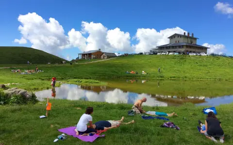 Persone rilassate sui prati verdi, vicino a un lago, sotto un cielo azzurro con nuvole bianche. Sembra un luogo ideale per i piani dell'avaro, godendosi la natura.